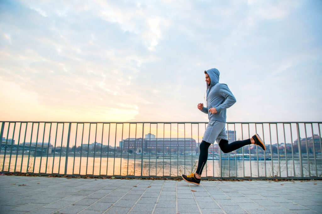 Person jogging in blue hoodie on paved waterfront path with metal railing and city skyline in background