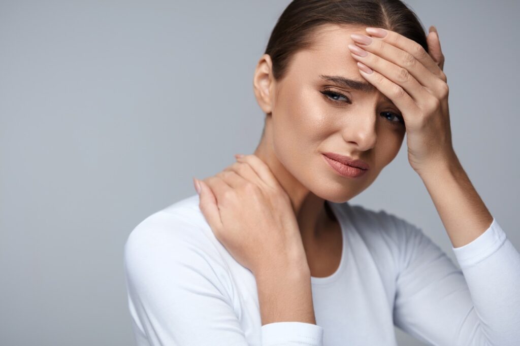 Woman in white shirt touching her forehead and neck, expressing discomfort or pain