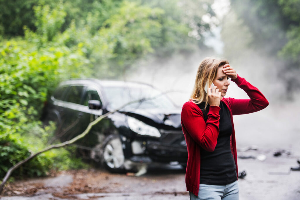 Woman in red cardigan standing beside damaged black car, holding hand to head while on phone call