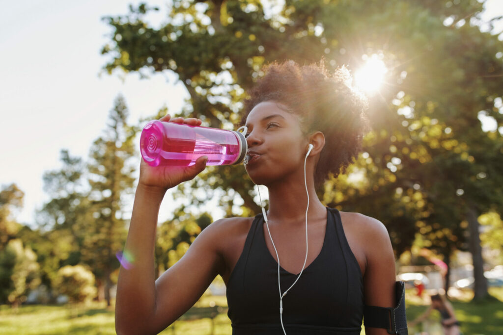 Woman in athletic wear drinking from pink water bottle outdoors in park with earbuds in