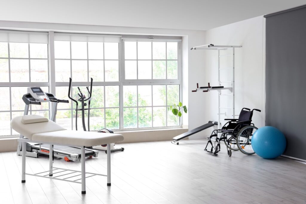 Modern physical therapy clinic with exercise equipment including treadmill, stationary bike, treatment table, wheelchair, blue exercise ball, and pull-up bar near large windows