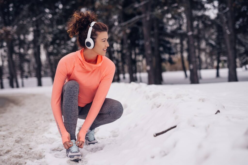 Woman in orange long-sleeve shirt and gray leggings stretching in snowy forest while wearing white headphones