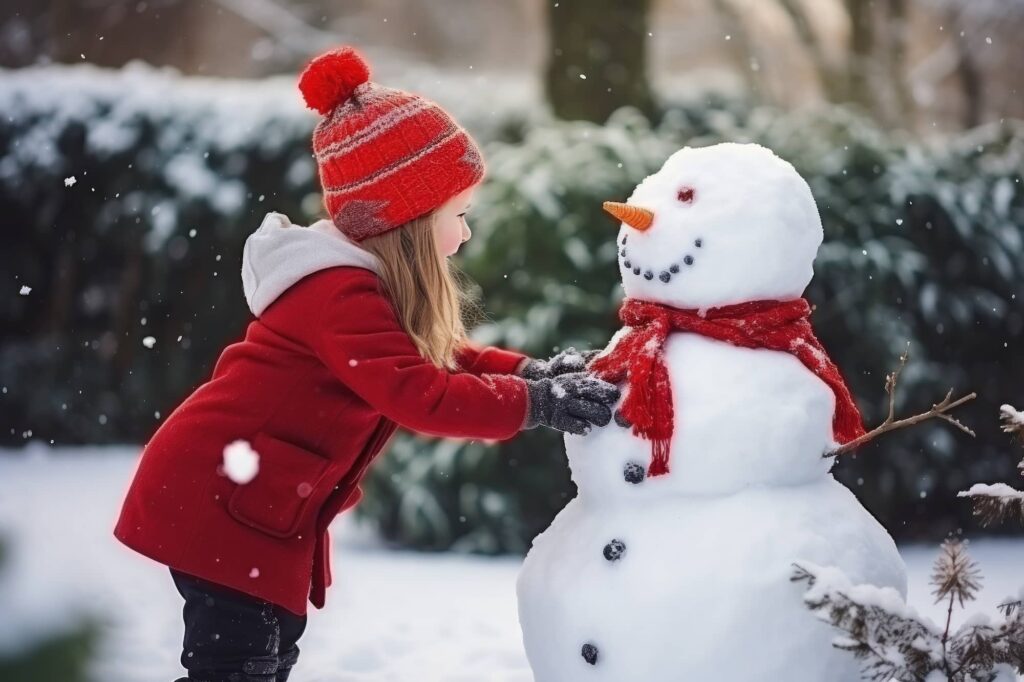 Child in red coat and knit hat touching snowman wearing red scarf in snowy winter garden