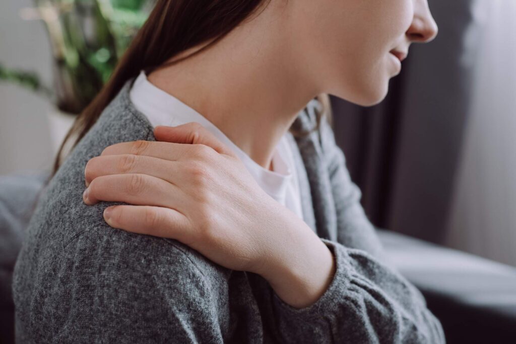 Woman in gray cardigan holding her shoulder, appearing to experience discomfort or stiffness