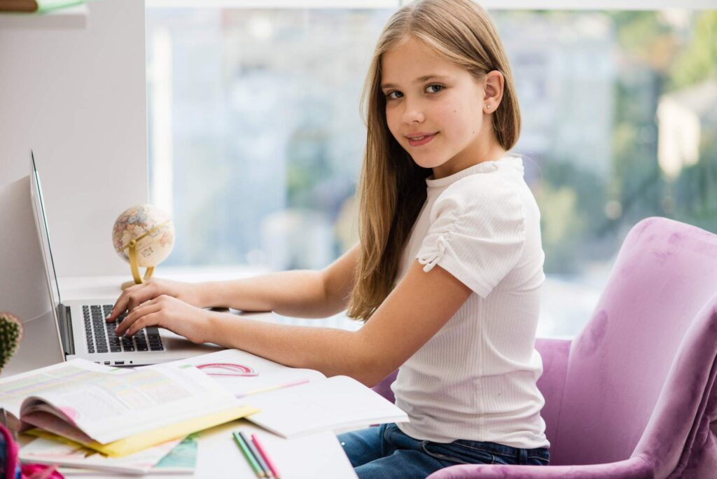 Young girl with blonde hair sitting at a desk with a laptop, globe, and study materials, looking over her shoulder at the camera with a slight smile
